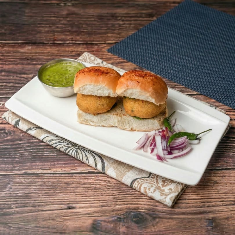 Two Mauli vada pavs on a white plate with green chutney, sliced red onions, and green chilies.