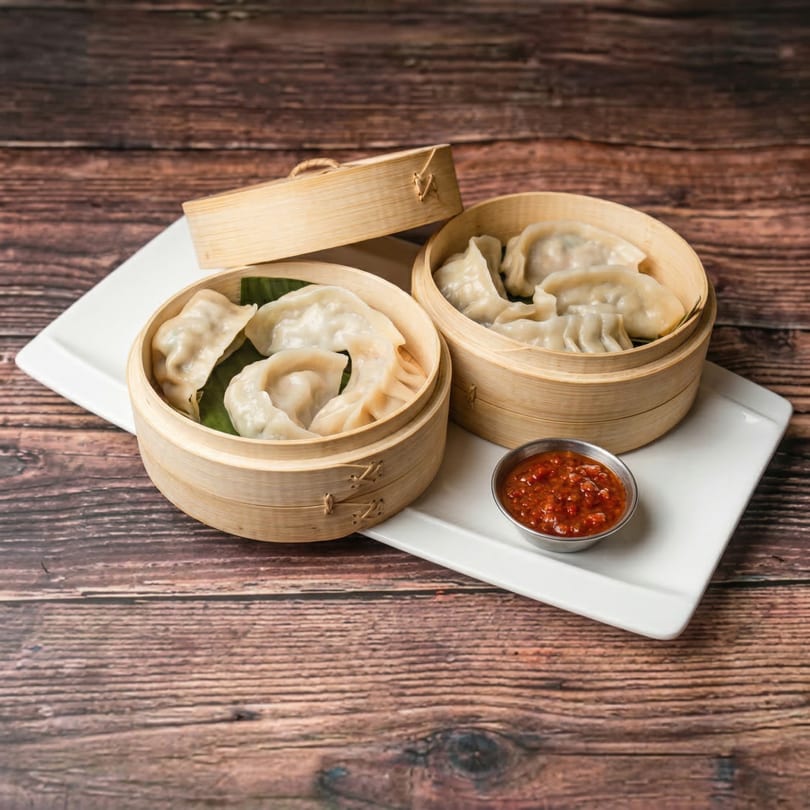 Two bamboo steamers filled with Mauli's Indian momos, served with a side of red chili sauce.