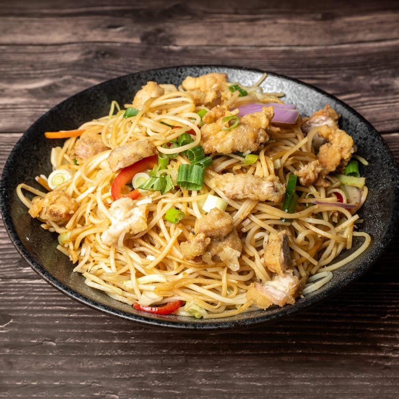 A black bowl filled with crispy chicken noodles, red peppers, and green onions on a wooden table.